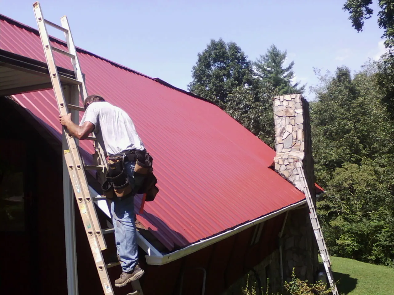 Roofer on a ladder beside a red metal roof and stone chimney — Soffit & Fascia Repair by Roofing Brothers in Oaklyn